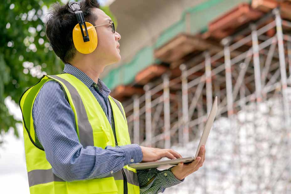 Safety officer wearing earmuffs logging noise risk monitoring on laptop